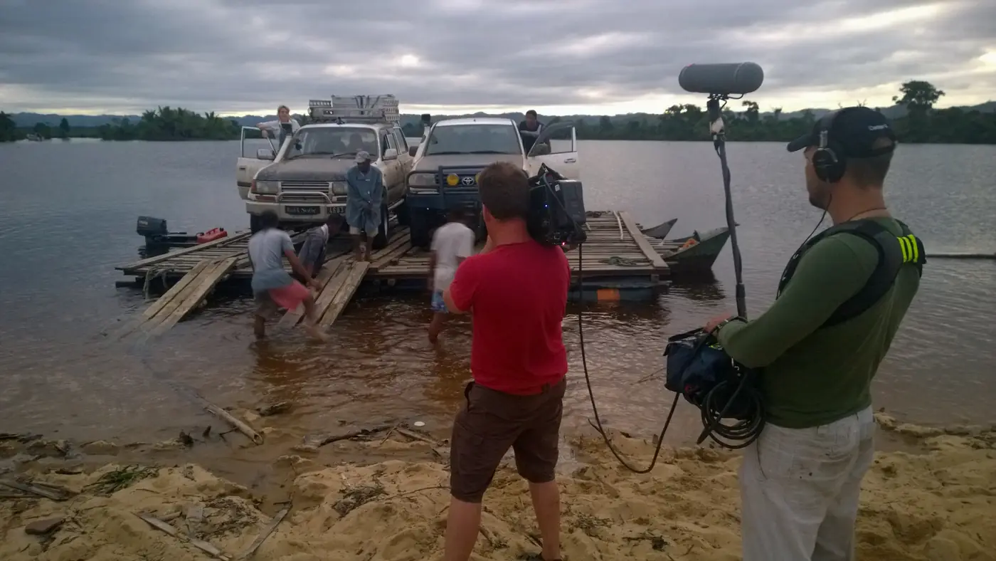Film crew recording 4x4 vehicles loaded onto a wooden river ferry in Madagascar